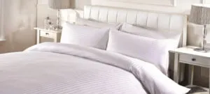 A neatly made bed with white striped bedding, matching pillows, and a padded white headboard, flanked by two bedside tables with lamps